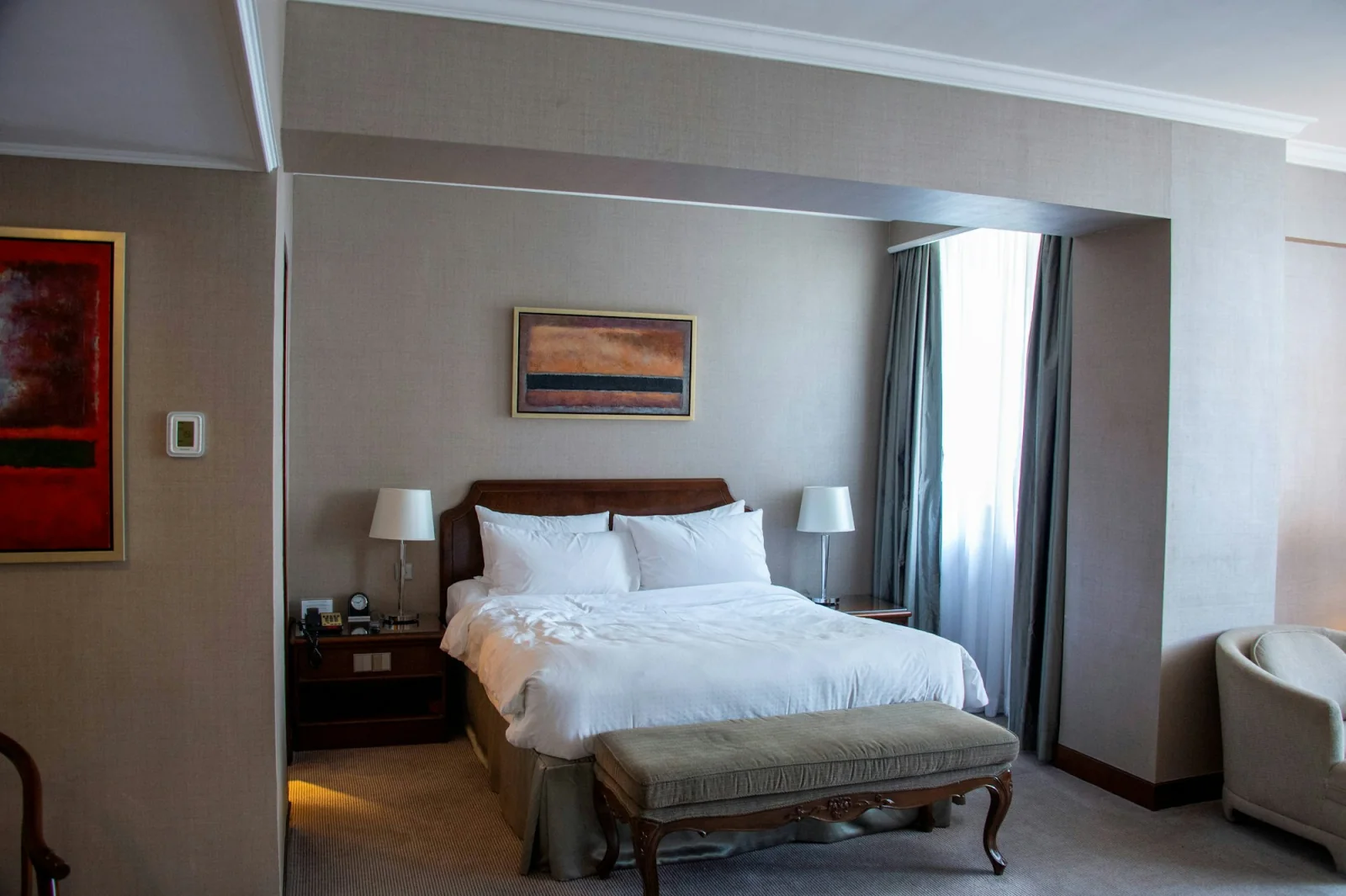 Hotel bedroom with white bedding, soft curtains and an upholstered bench.