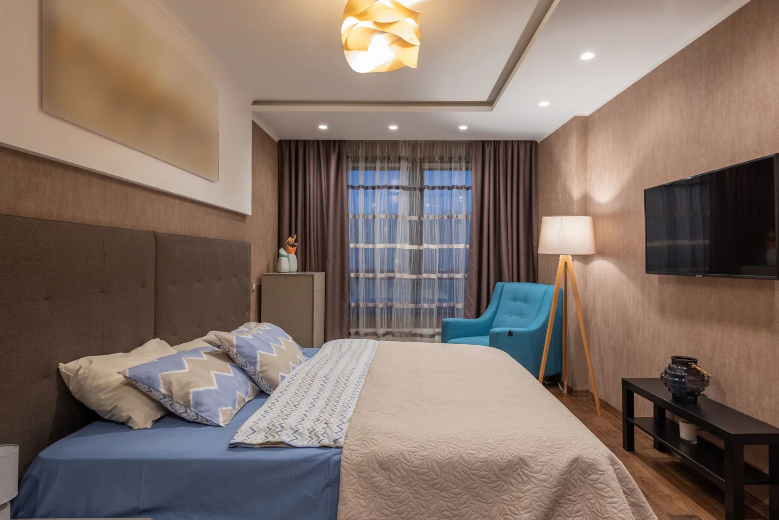 Hotel bedroom with layered bedding, a blue chair and tall curtains.